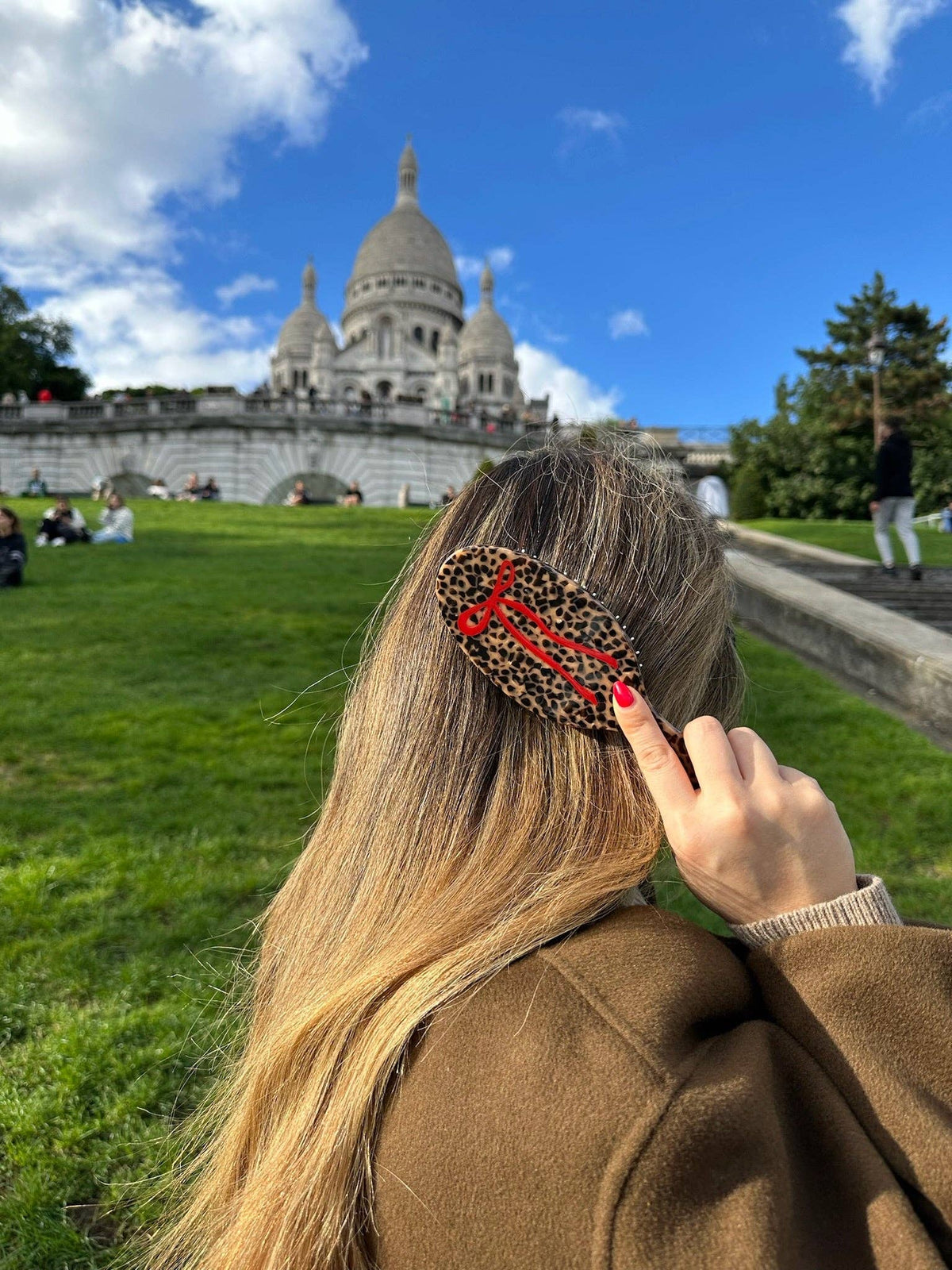 Hand-painted leopard print hair bow brush with red cord, held against blue sky and cathedral backdrop.