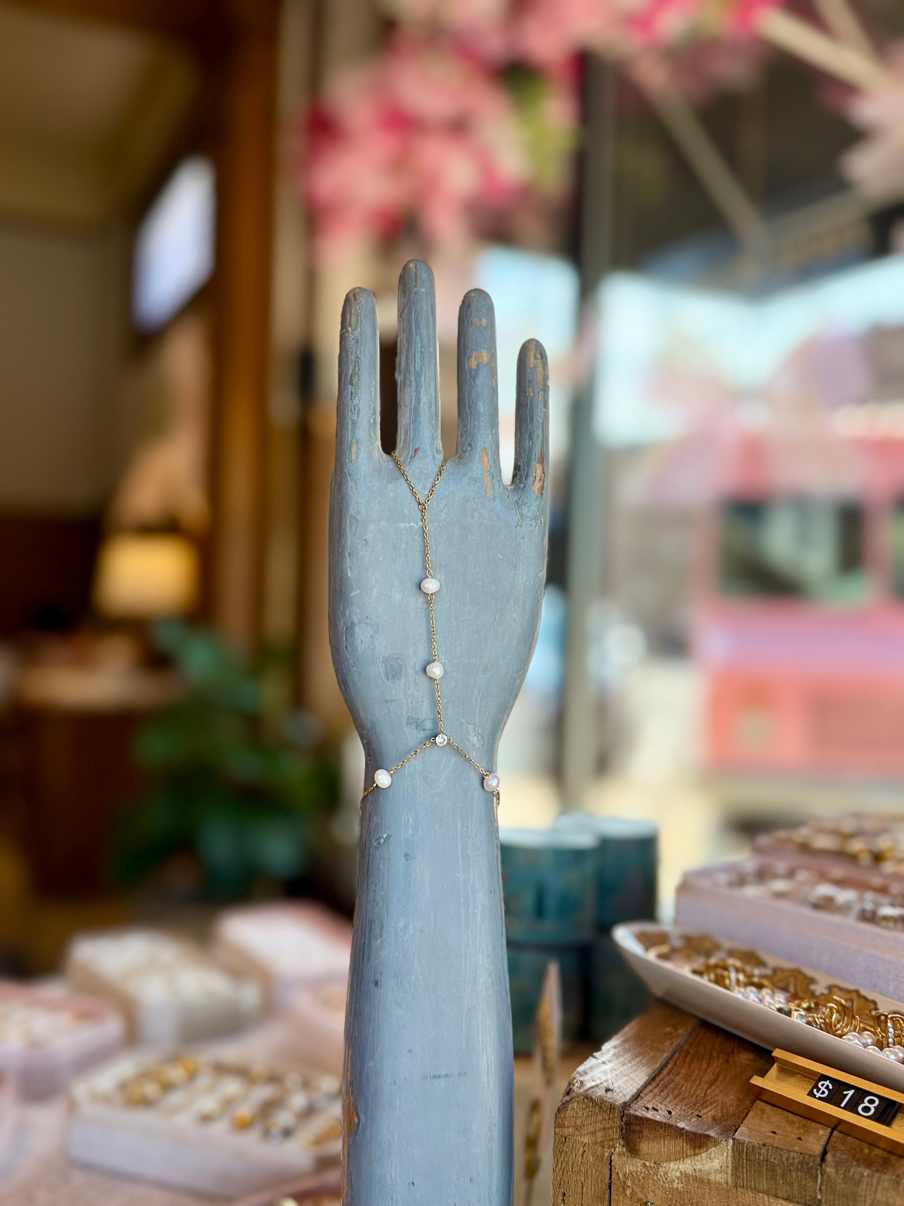 Pearlie Hand Chain bracelet on a weathered blue display hand, gold chain with tiny pearl beads, jewelry shop backdrop.