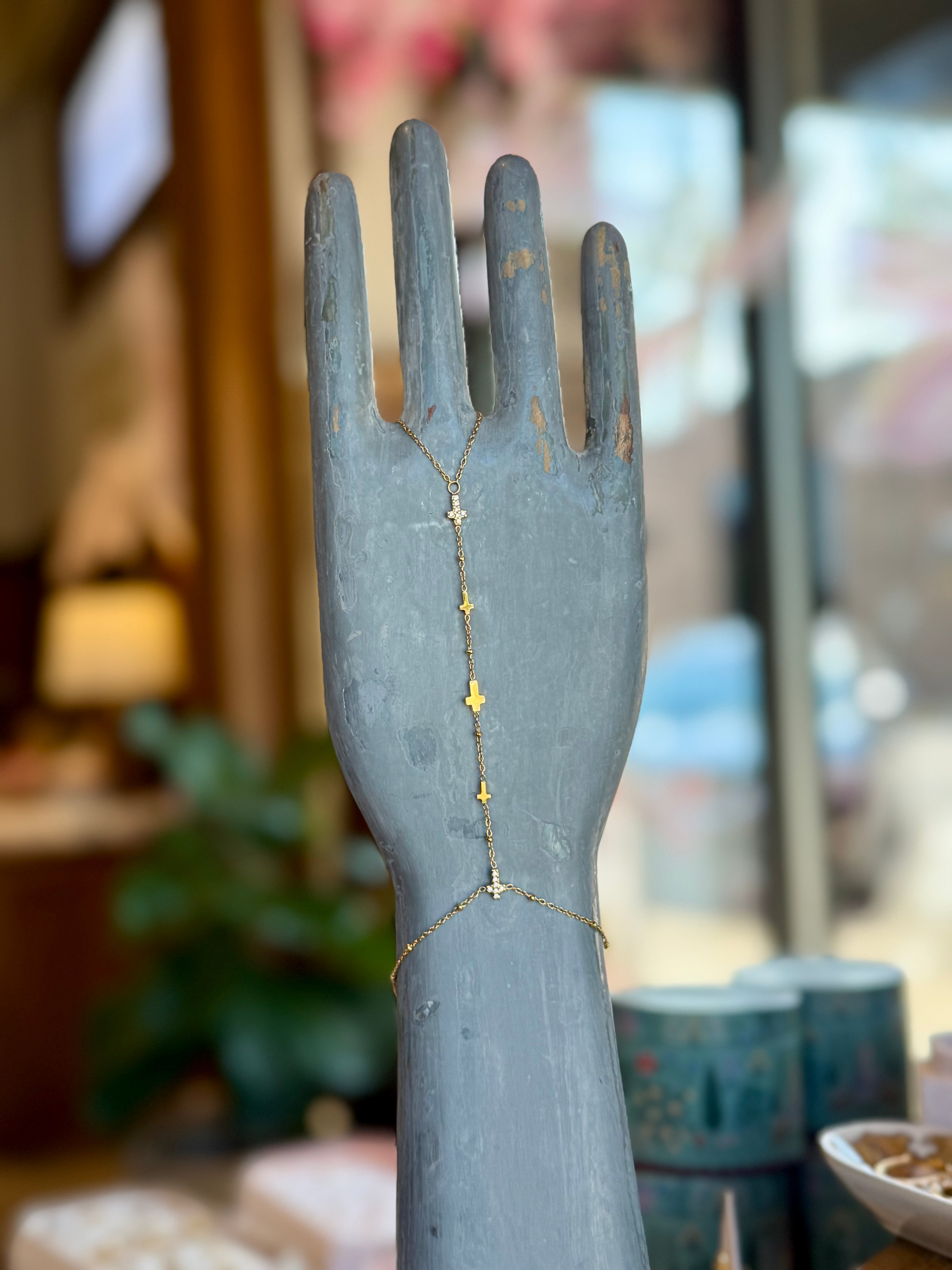 Sacred Hand Chain bracelet in gold-tone metal with cross charms, draped on a distressed blue-gray hand display.